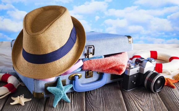 Packed suitcase for summer holidays - hat, towel, flip flops and photo camera on wooden and beach background